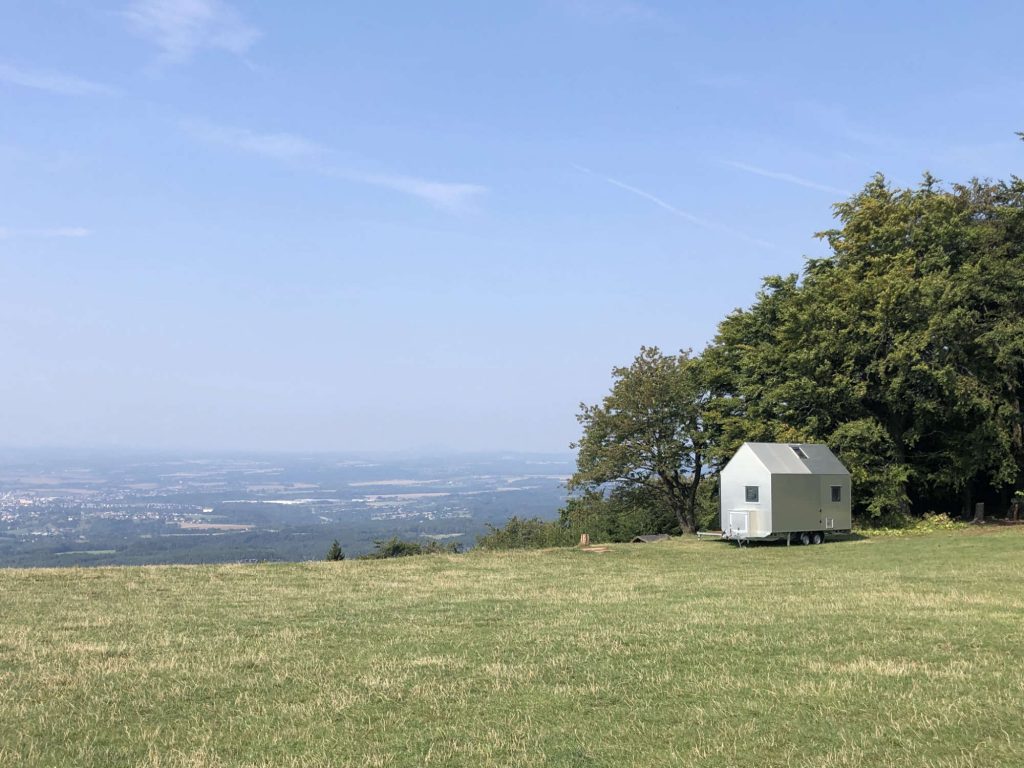 Tiny house na podvozku umístěný na vrcholu kopce s výhledem na širokou krajinu. Dům má moderní hliníkovou fasádu a solární panely na střeše.