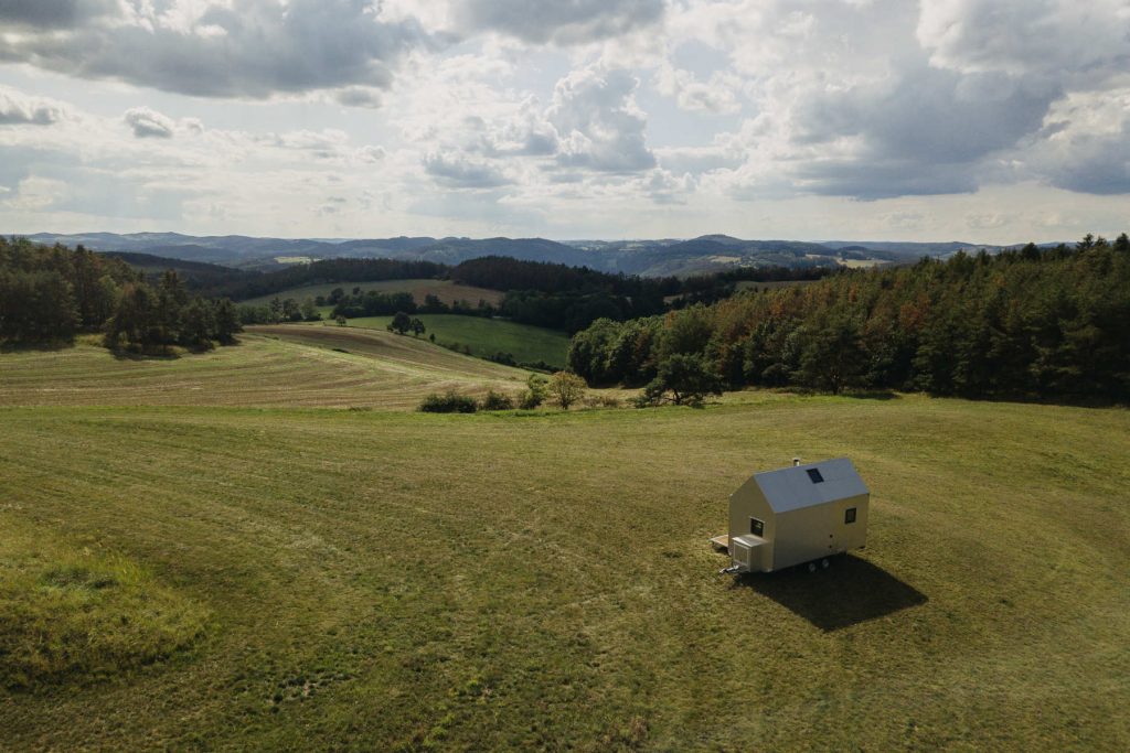 Tiny house na podvozku stojí na rozlehlé louce obklopené kopcovitou krajinou. Moderní dům s jednoduchým designem ladí s přírodním prostředím, podtrženým horami v pozadí.