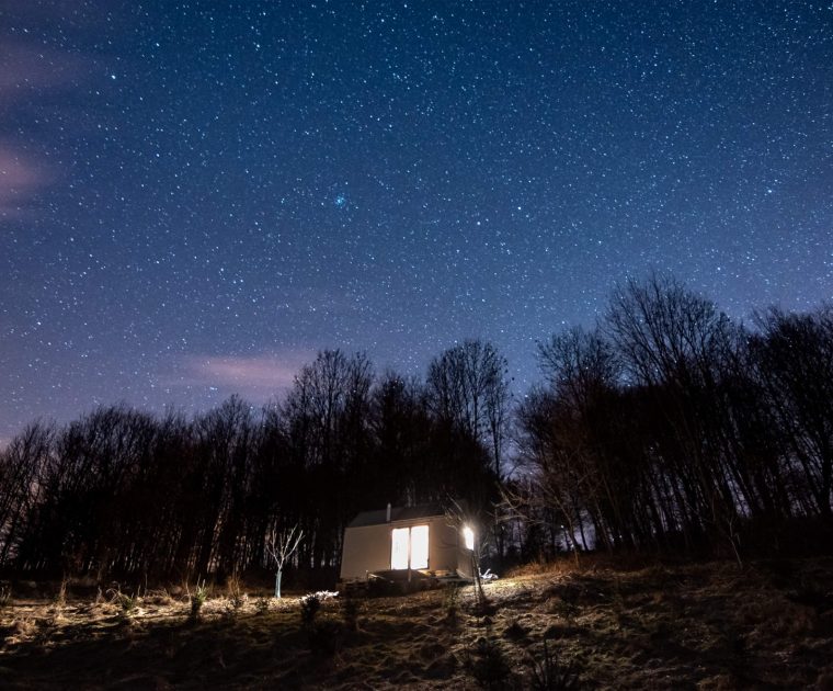 Osvětlený mobilní dům v noční krajině pod hvězdnou oblohou, zasazený na okraji lesa, ideální pro off-grid bydlení a únik do přírody.